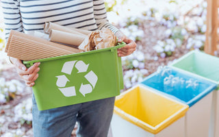 Woman holding a green recycling pin full of cardboard is standing frot of three color-coded waste bins.