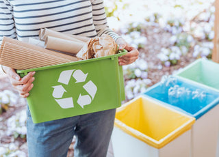 Woman holding a green recycling pin full of cardboard is standing frot of three color-coded waste bins.