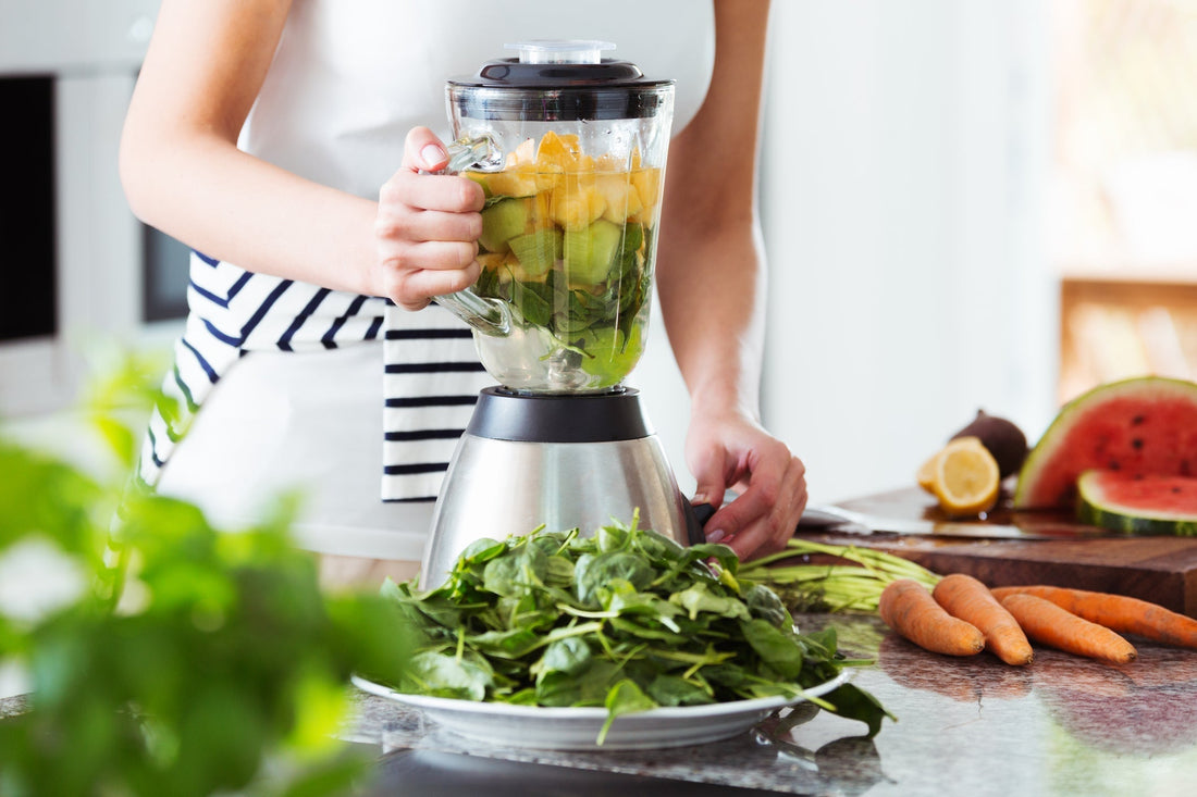 Woman preparing a healthy smoothie with greens, citrus, and cucumber in the kitchen with carrots sitting next to the blender on the countertop. 