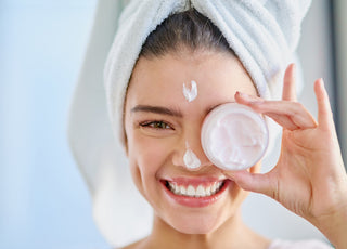 Young woman with her hair wrapped up in a white towel holding an open jar of moisturizer over her eye.