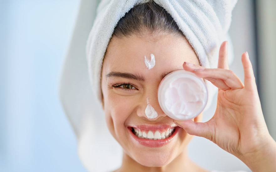 Young woman with her hair wrapped up in a white towel holding an open jar of moisturizer over her eye.