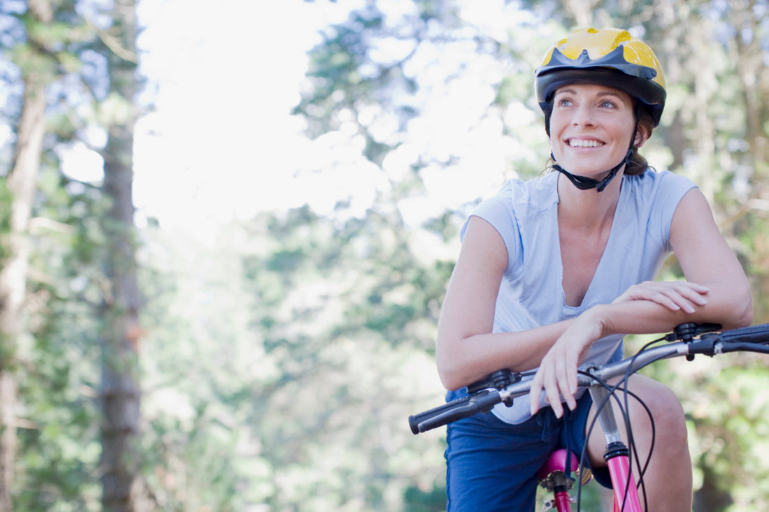 Woman wearing a yellow and black helmet and blue workout gear is sitting on a pink bike and looking off into the distance.