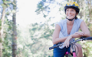 Woman wearing a yellow and black helmet and blue workout gear is sitting on a pink bike and looking off into the distance.