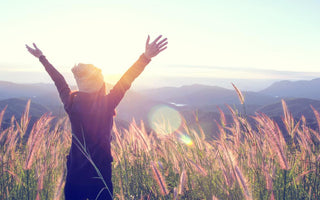 Young women standing in a field of tall grass holding her arms up in the air as she takes in the mountain view. 