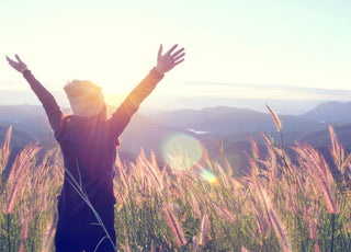 Young women standing in a field of tall grass holding her arms up in the air as she takes in the mountain view. 