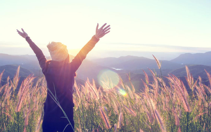 Young women standing in a field of tall grass holding her arms up in the air as she takes in the mountain view. 