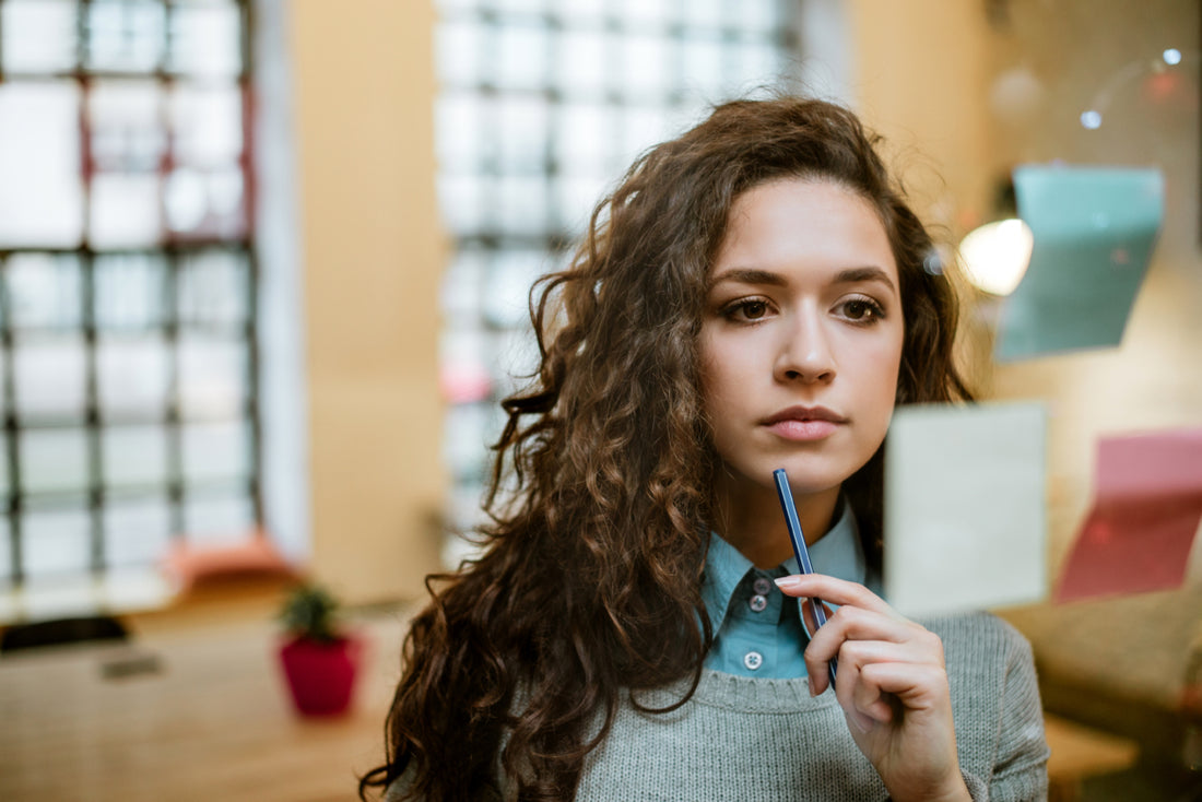 Business woman standing in an office holding a pen to her chin as she look as a sticky note on a glass wall.
