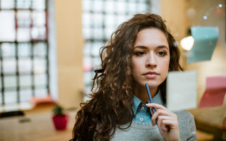 Business woman standing in an office holding a pen to her chin as she look as a sticky note on a glass wall.