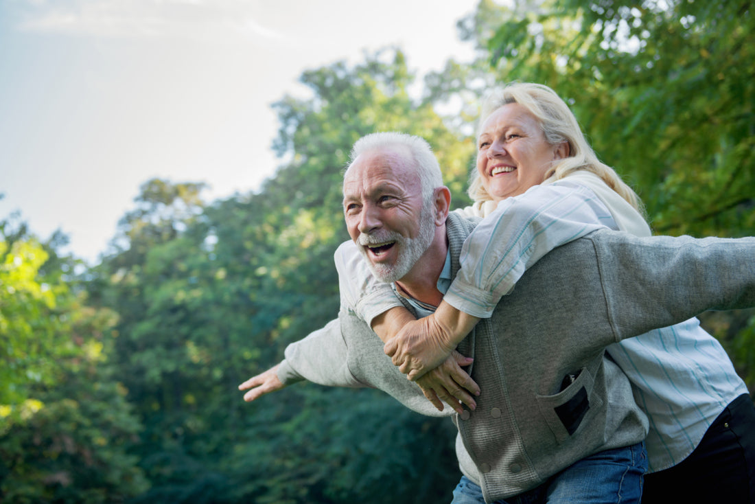 An elderly man bent over slightly at the waist with his arms out to the side while an elderly woman leans over his back and wraps her arms around his neck.