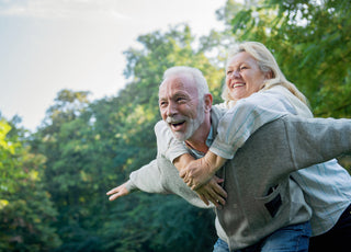 An elderly man bent over slightly at the waist with his arms out to the side while an elderly woman leans over his back and wraps her arms around his neck.