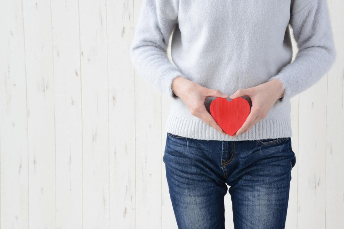 Female in jeans and grey sweater holding a red wooden heart over her lower stomach.