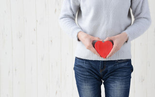 Female in jeans and grey sweater holding a red wooden heart over her lower stomach.