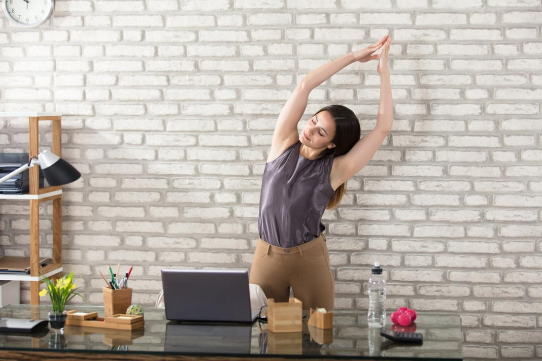 Young adult woman standing at her desk stretching with her arms above her head.