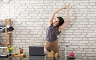 Young adult woman standing at her desk stretching with her arms above her head.