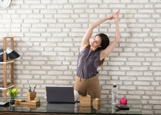 Young adult woman standing at her desk stretching with her arms above her head.