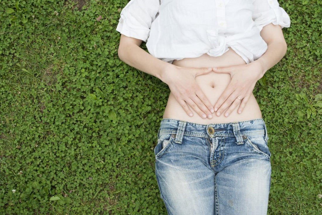 Women in a white shirt and jeans laying on a patch of bright green clover holding her hands in a triangle shape over her belly button. 