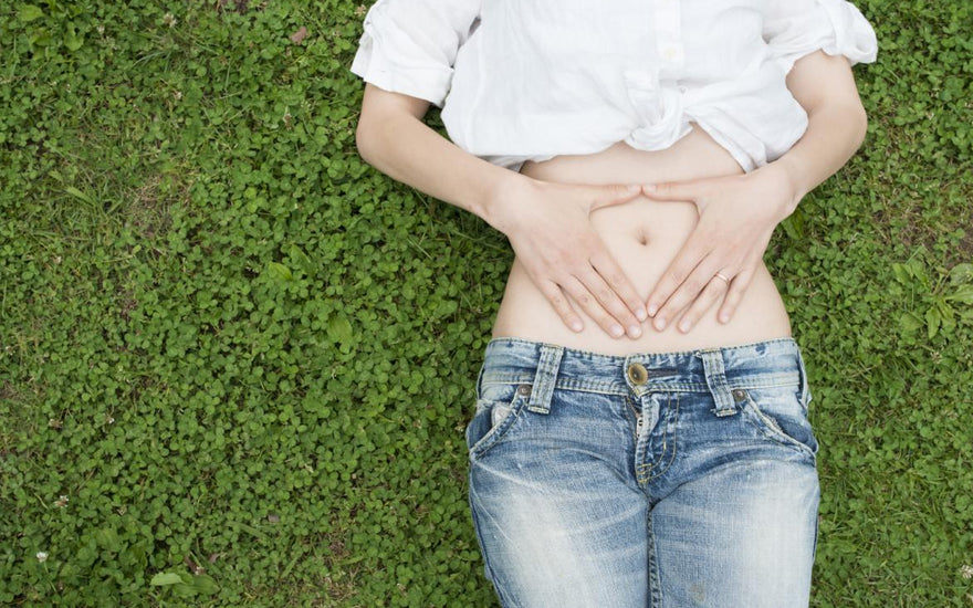 Women in a white shirt and jeans laying on a patch of bright green clover holding her hands in a triangle shape over her belly button. 