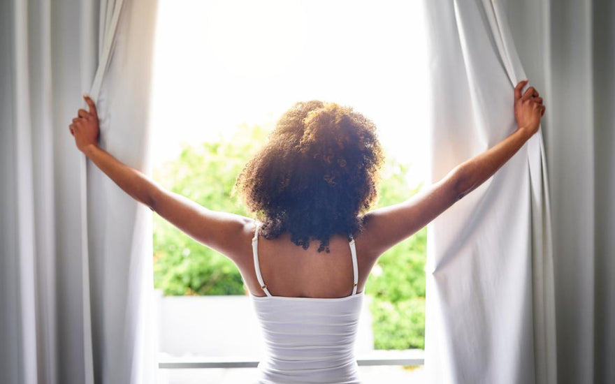 Women in a white tank top standing in front of the window while she opens the curtains and looks out at the morning view.