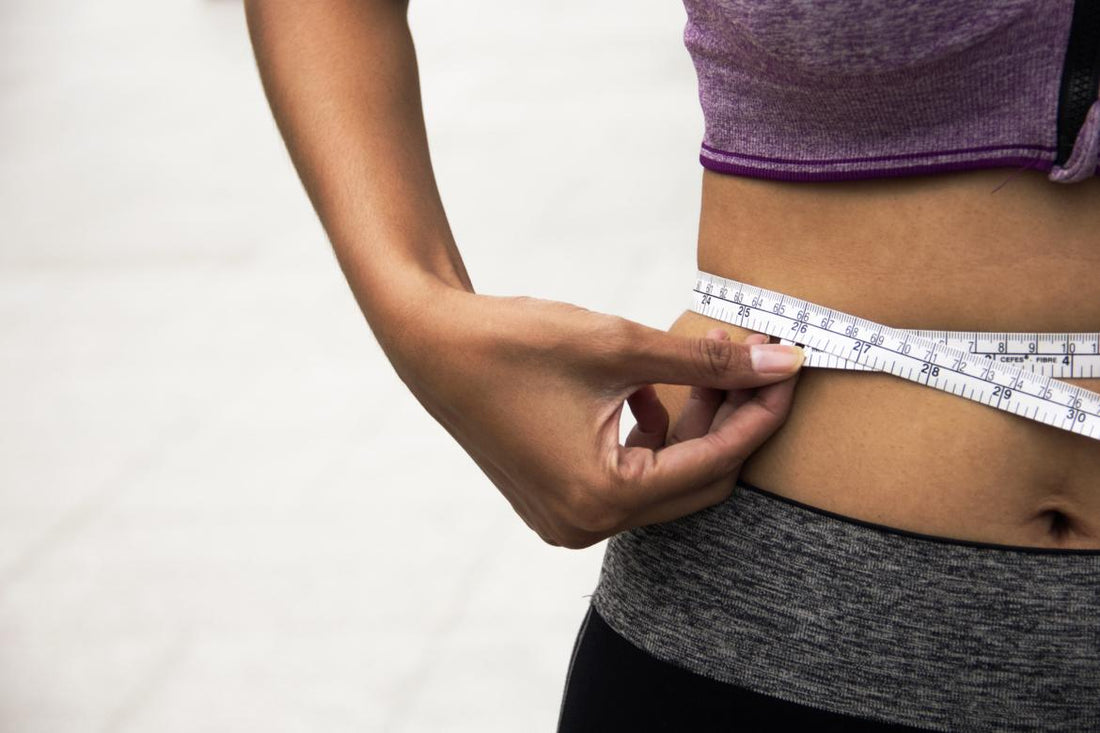 Woman in workout gear using a cloth tape measure to measure her waist.