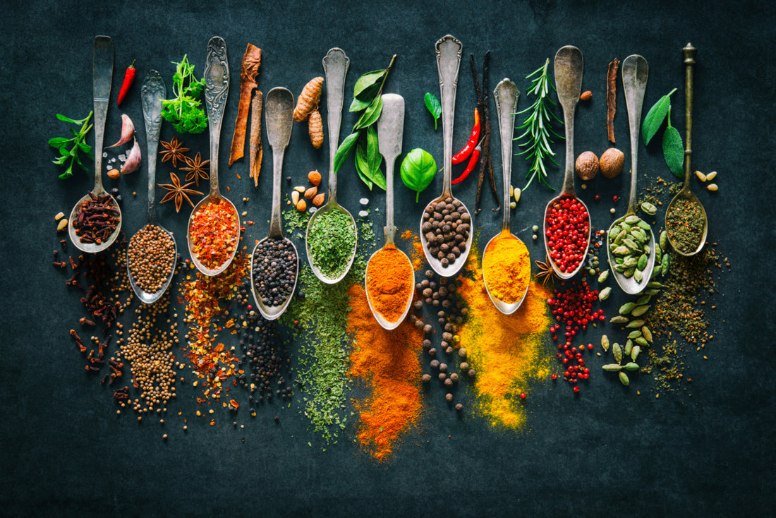 Eleven metal spoons on a black background each filled with a different herb or spice and surrounded by the foods used to create those powders.