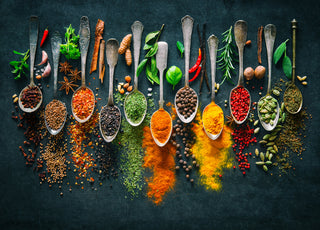 Eleven metal spoons on a black background each filled with a different herb or spice and surrounded by the foods used to create those powders.