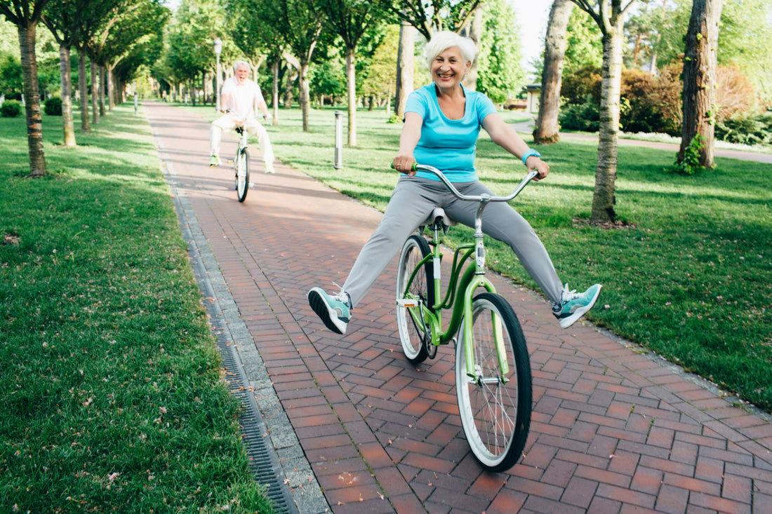 An elderly woman riding a green bike down a brick path is smiling and holding her feet in the air. There is an elderly man right behind her participating in the game action.