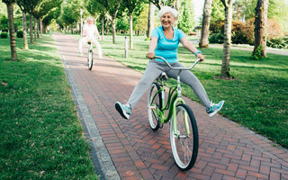 An elderly woman riding a green bike down a brick path is smiling and holding her feet in the air. There is an elderly man right behind her participating in the game action.