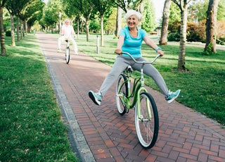 An elderly woman riding a green bike down a brick path is smiling and holding her feet in the air. There is an elderly man right behind her participating in the game action.