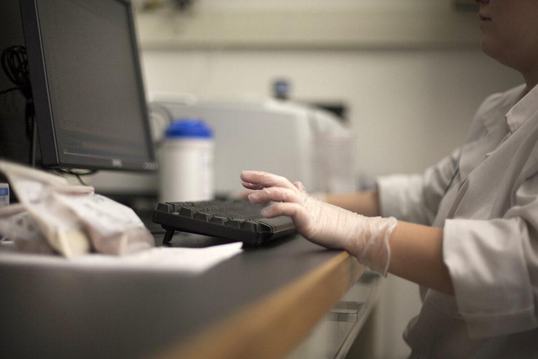 Scientist in a white goat and gloves typing on a desktop computer in a lab. 