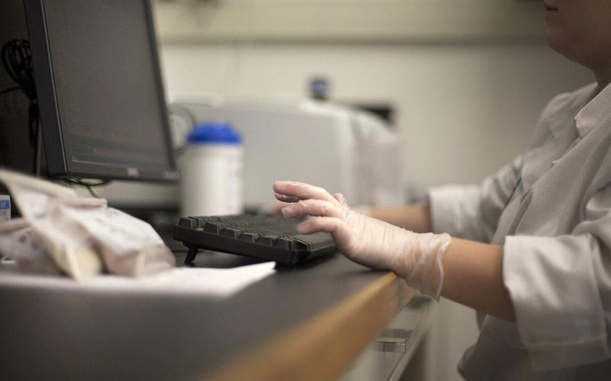 Scientist in a white goat and gloves typing on a desktop computer in a lab. 