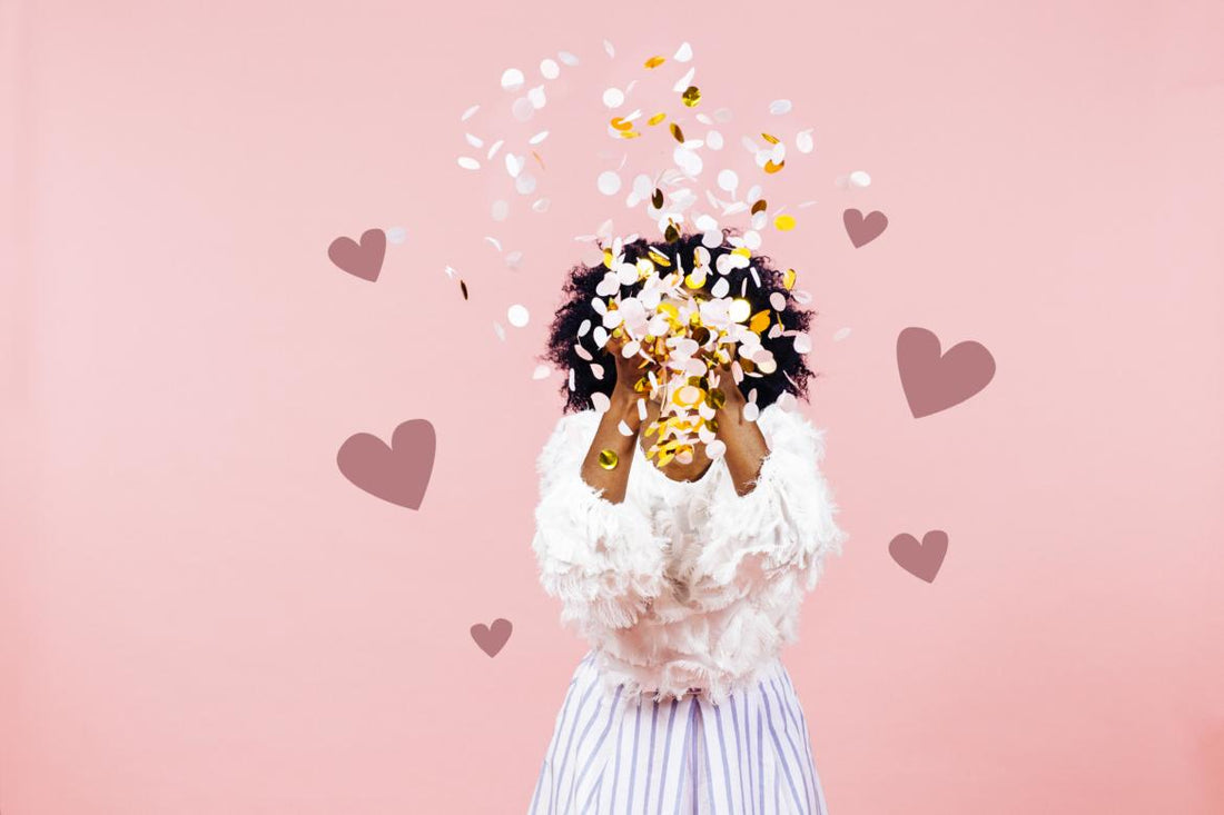 Woman standing in front of a pink background throwing confetti in front of her face.