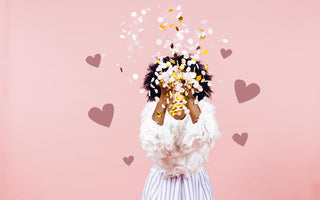 Woman standing in front of a pink background throwing confetti in front of her face.