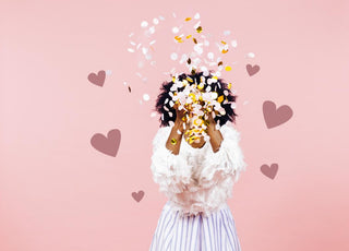 Woman standing in front of a pink background throwing confetti in front of her face.