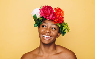 Woman of color with a flower headband on her head standing and smiling in front of a yellow background.