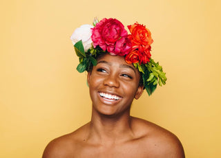 Woman of color with a flower headband on her head standing and smiling in front of a yellow background.