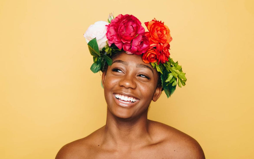 Woman of color with a flower headband on her head standing and smiling in front of a yellow background.