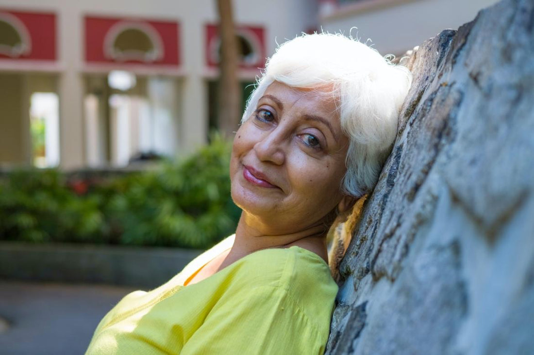 Older woman with short white hair leaning against a rock wall in a city center.