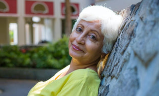 Older woman with short white hair leaning against a rock wall in a city center.