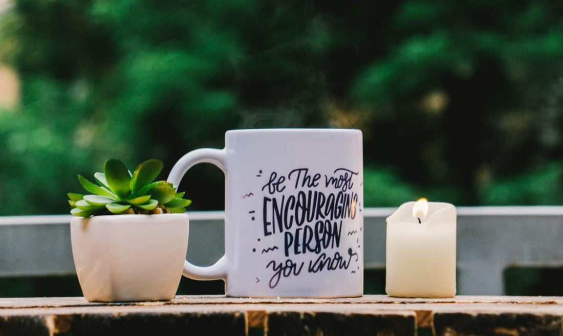 White coffee mug with black writing sitting on an outdoor table next to a candle and a small potted plant.