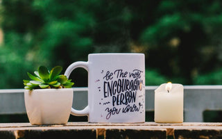 White coffee mug with black writing sitting on an outdoor table next to a candle and a small potted plant.