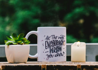 White coffee mug with black writing sitting on an outdoor table next to a candle and a small potted plant.