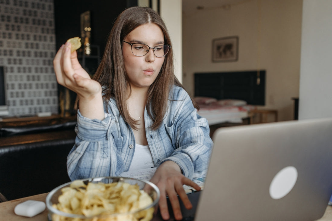 Woman in a blue plaid shirt and glasses working on her laptop at a table while snacking on potato chips.