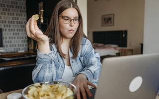 Woman in a blue plaid shirt and glasses working on her laptop at a table while snacking on potato chips.