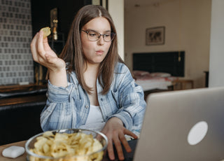 Woman in a blue plaid shirt and glasses working on her laptop at a table while snacking on potato chips.