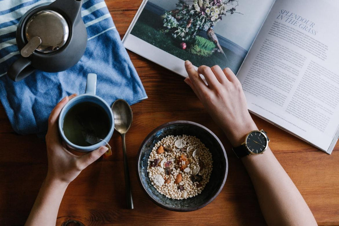 Top down view of a pair of hands holding a mug full of tea and flipping through a book on a wooden table that also has bowl of oatmeal on it. 