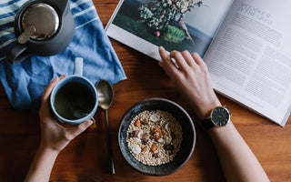 Top down view of a pair of hands holding a mug full of tea and flipping through a book on a wooden table that also has bowl of oatmeal on it. 