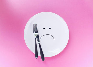 A plate with a sad face drawn on it sitting on a pink cloth with a fork and knife sitting on top of the plate.