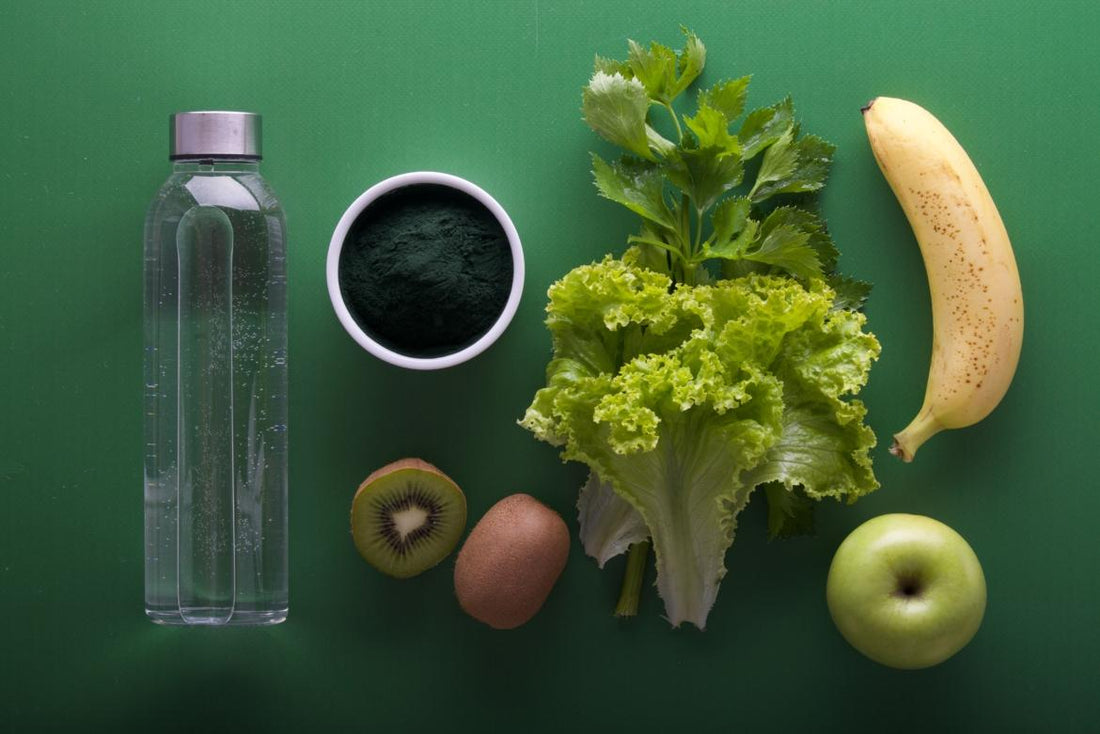 Flat lay of healthy foods including lettuce, a banana, kiwi, and algae powder next to a bottle of water. 