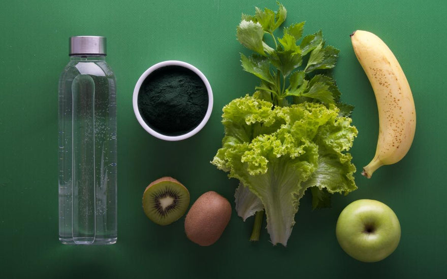 Flat lay of healthy foods including lettuce, a banana, kiwi, and algae powder next to a bottle of water. 